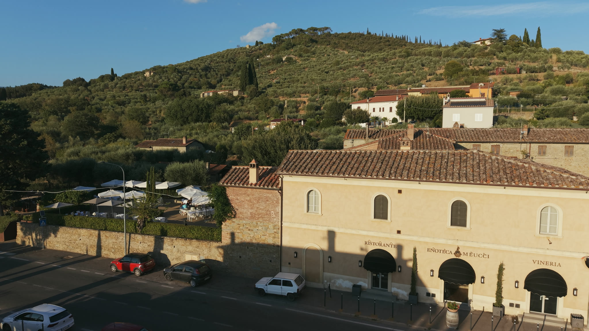 Il casale dell'Enoteca Meucci visto dalla strada: il giardino con gli ombrelloni a sinistra, le tre arcate del ristorante, dell'enoteca e della vineria, la collina di olivi alle spalle.