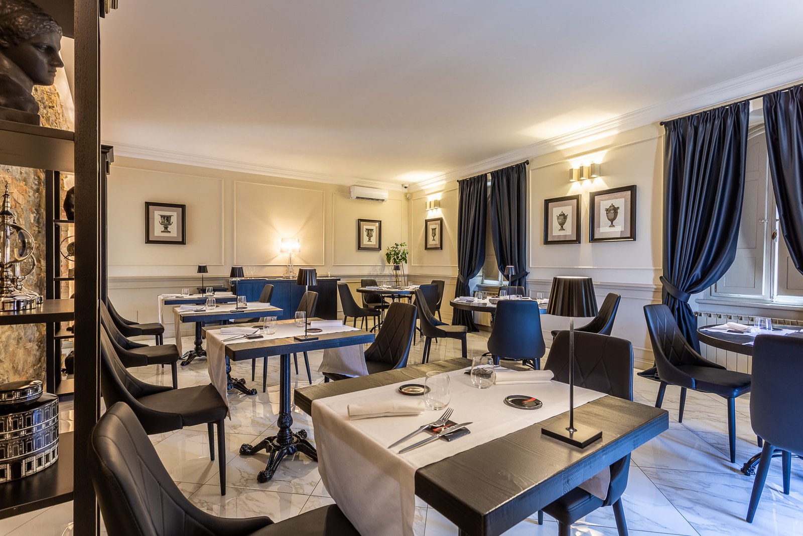La sala al piano di sopra: tavoli con la tovaglia bianca, boiserie chiara, tende di velluto scuro e stampe incorniciate alle pareti